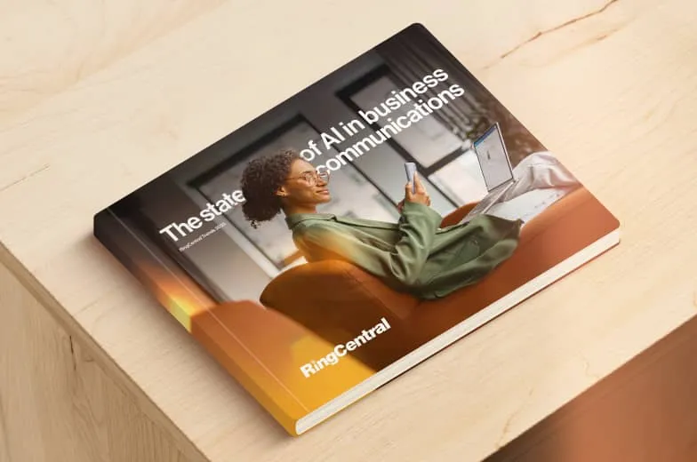 A view of the 'The state of AI in business communications' report by RingCentral on a wooden table, featuring a woman on the cover using a smartphone and laptop.